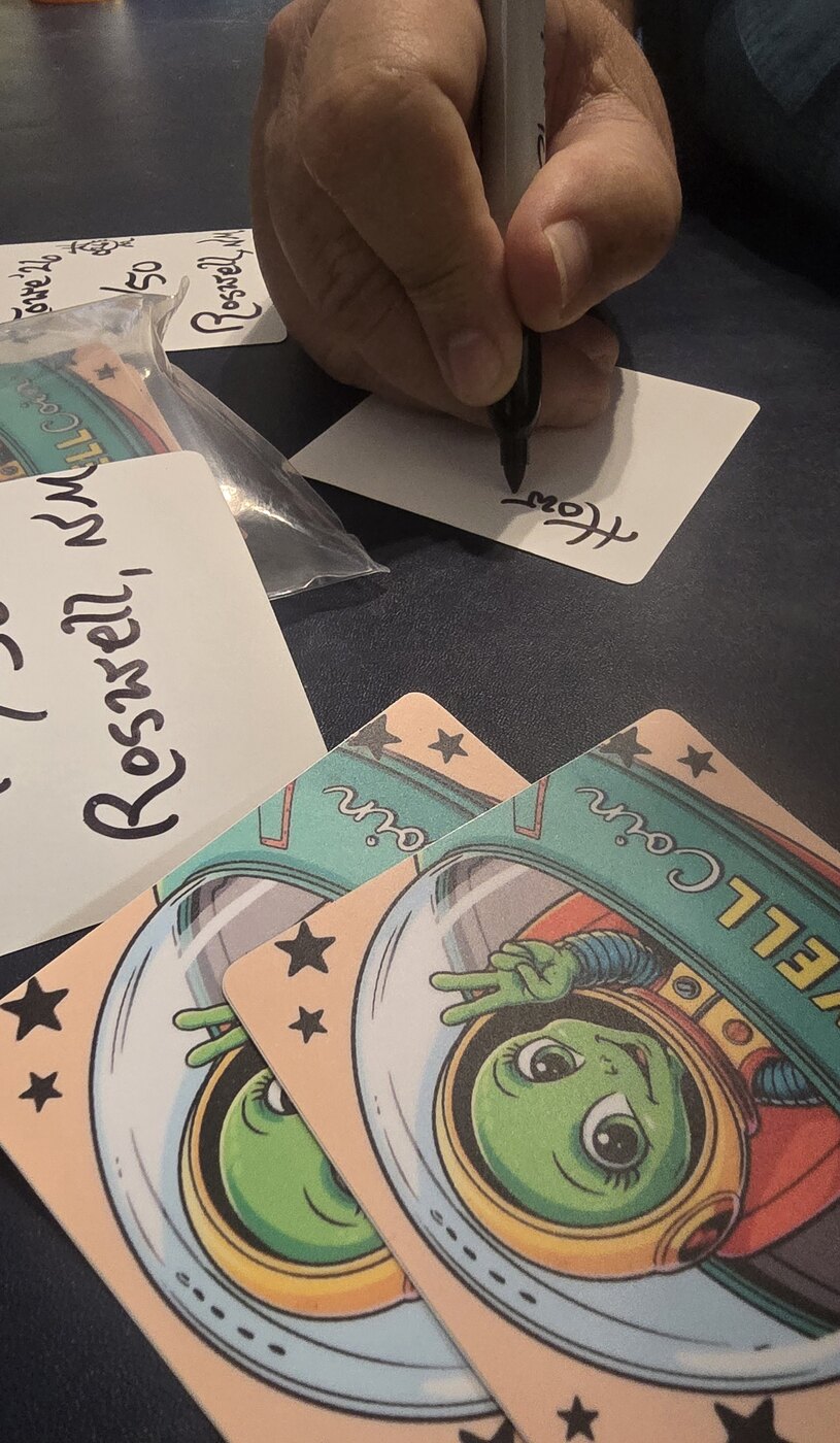 The creator's hand signing and numbering RoswellCoin stickers; no face is shown